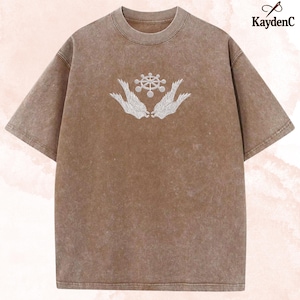 May include: Brown t-shirt with a crew neck and short sleeves. The shirt features a white embroidered design of a wheel-like symbol held by two stylized wings. The shirt has a vintage, washed appearance.