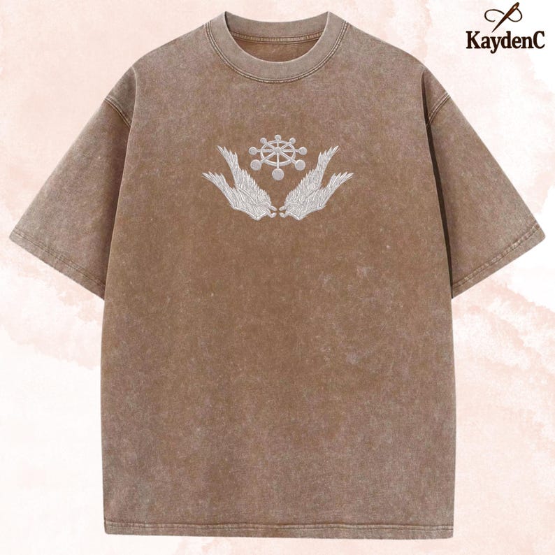 May include: Brown t-shirt with a crew neck and short sleeves. The shirt features a white embroidered design of a wheel-like symbol held by two stylized wings. The shirt has a vintage, washed appearance.