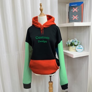 May include: A color-block hoodie with a black body, orange hood and pocket, and mint green sleeves. The text "Custom Design" is printed in green on the front. The hoodie is displayed on a mannequin.