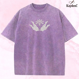 May include: A lilac-coloured t-shirt with a faded, textured finish. It has an embroidered design of a central circular element flanked by wing-like shapes. The brand name "KaydenC" is visible in the upper right corner.
