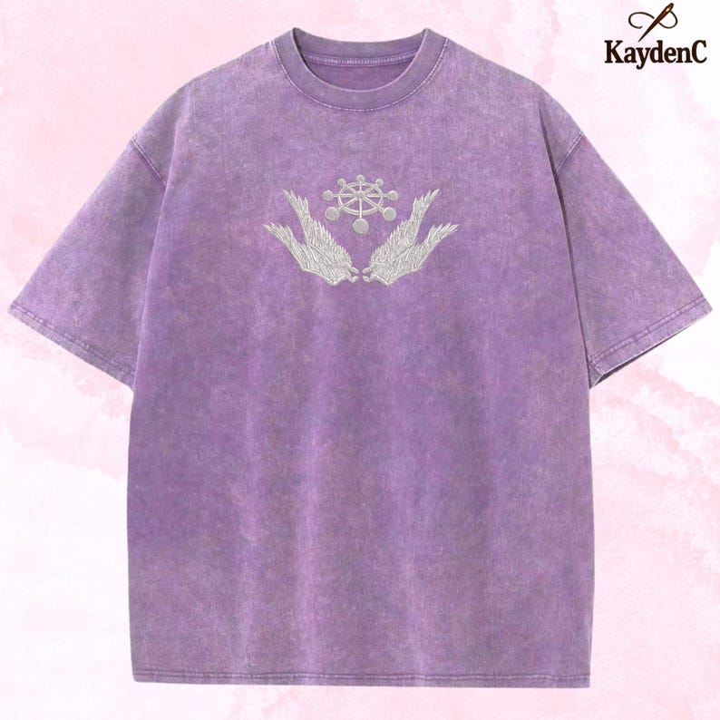 May include: A lilac-coloured t-shirt with a faded, textured finish. It has an embroidered design of a central circular element flanked by wing-like shapes. The brand name "KaydenC" is visible in the upper right corner.