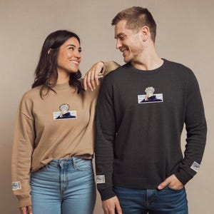 Pode incluir: Um casal vestindo su&eacute;teres de manga comprida combinando com um gr&aacute;fico de personagem de desenho animado na frente. Os su&eacute;teres s&atilde;o bege e cinza escuro. O casal est&aacute; olhando um para o outro e sorrindo.