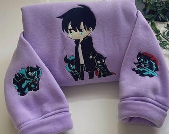 Puede incluir: Una sudadera de color morado claro con un personaje de estilo anime de pelo negro bordado en el centro. Las mangas muestran figuras bordadas adicionales, incluyendo una criatura con cuernos a la izquierda y un guerrero oscuro a la derecha.