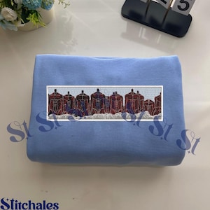May include: A light blue sweatshirt with an embroidered rectangular design. The design features several muscular figures in shades of red and brown, with a blue and white wave pattern at the bottom. The sweatshirt is folded neatly on a white surface.