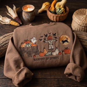 Peut inclure: Un sweat-shirt marron orné d'une broderie représentant une charrette en bois remplie de citrouilles, un fantôme, un épouvantail à tête de citrouille, des corbeaux en vol et un chat noir devant une pleine lune, avec le texte Harvest of Spirits.