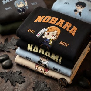 Może przedstawiać: Stos złożonych bluz z wyhaftowanymi postaciami w stylu anime i tekstem. G&oacute;rna czarna bluza zawiera napis NOBARA oraz tekst EST. 2017. Poniżej znajdują się bluzy w kolorze czarnym, jasnoniebieskim i beżowym z r&oacute;żnymi wzorami postaci.