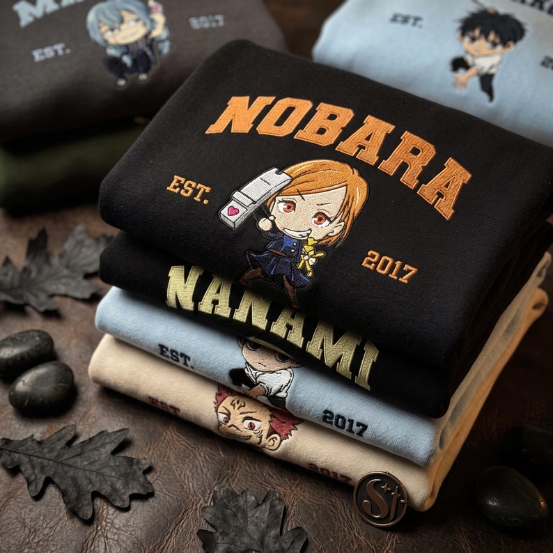Może przedstawiać: Stos złożonych bluz z wyhaftowanymi postaciami w stylu anime i tekstem. G&oacute;rna czarna bluza zawiera napis NOBARA oraz tekst EST. 2017. Poniżej znajdują się bluzy w kolorze czarnym, jasnoniebieskim i beżowym z r&oacute;żnymi wzorami postaci.