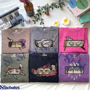 May include: Six mineral-washed crewneck t-shirts in brown, grey, pink, olive green, black, and purple. Each shirt features a rectangular embroidered graphic of anime-style character eyes surrounded by thematic elements like swords, bandages, and skulls.