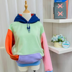 May include: A multi-coloured hoodie with a light green body, orange left sleeve, pink right sleeve, and a blue hood. The hoodie has a purple pocket and a blue trim around the bottom.