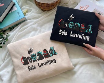 May include: Two sweatshirts, one black and one beige, each featuring an embroidered graphic of various stylized characters and the text Solo Leveling in a gothic font. The designs include small figures with wings, horns, and colorful accents.