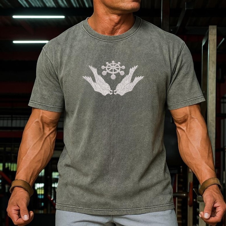 May include: A grey short-sleeved t-shirt with a white graphic design of a stylized emblem with wings. The shirt has a vintage, washed appearance. The model is wearing a brown leather bracelet on each wrist.