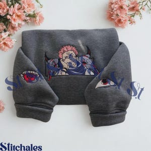 Könnte beinhalten: Ein dunkelgraues Sweatshirt mit einer Stickerei eines Anime-Charakters mit rosa Haaren und dunklen Markierungen auf der Brust. Die Ärmel sind mit gestickten Augenmotiven verziert, eines mit roter Iris und das andere mit einem weißen Augapfel.