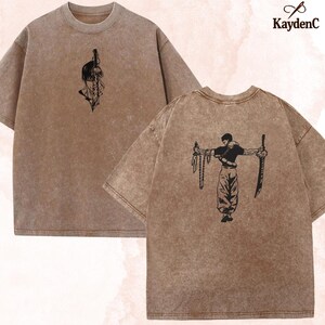 Puede incluir: Camisetas marrones con un aspecto descolorido y vintage. Una camiseta presenta un gr&aacute;fico negro de una persona con una espada en la espalda y una cara con una espada en la parte delantera. La marca "KaydenC" est&aacute; en la esquina superior derecha.