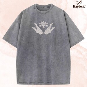 May include: Grey crew neck t-shirt with short sleeves. It has a white embroidered design of a wheel-like symbol held by two stylized wings. The brand name "KaydenC" is printed in the upper right corner.