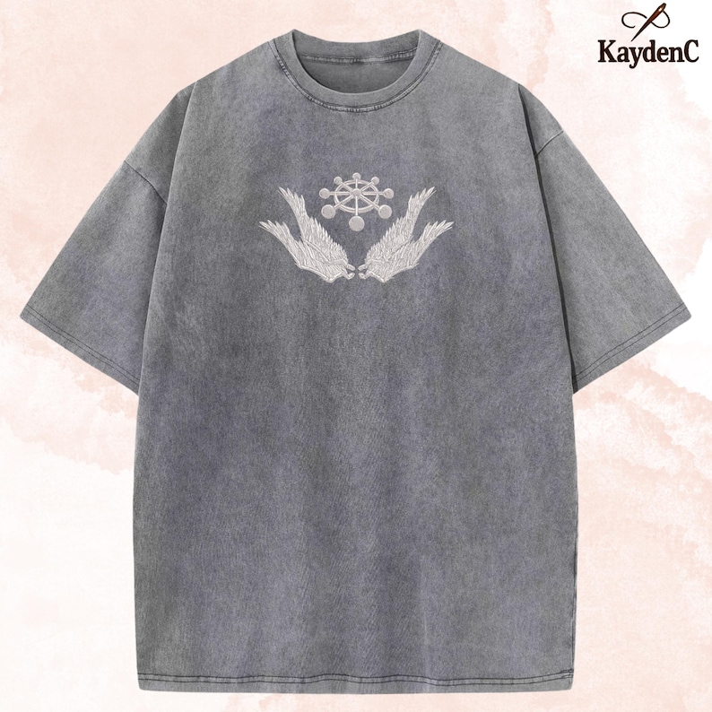 May include: Grey crew neck t-shirt with short sleeves. It has a white embroidered design of a wheel-like symbol held by two stylized wings. The brand name "KaydenC" is printed in the upper right corner.