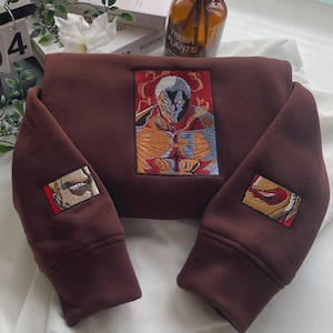 Op de afbeelding: Een donkerbruin sweatshirt met lange mouwen en een grote rechthoekige geborduurde afbeelding op de borst van een gespierd personage met grijs haar en rode accenten. Kleinere rechthoekige geborduurde patches bevinden zich op elke mouw.