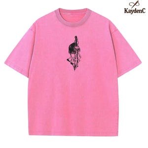 Puede incluir: Una camiseta rosa con cuello redondo y mangas cortas. La camiseta presenta un gr&aacute;fico negro de una persona con una espada. La marca "KaydenC" est&aacute; en la esquina superior derecha.