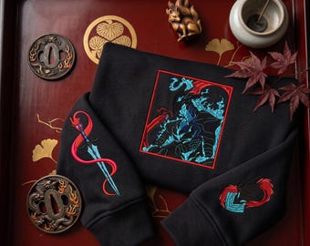 May include: A black sweatshirt featuring a central rectangular embroidery of a dark figure surrounded by blue flames. The left sleeve displays a blue sword wrapped in a red ribbon, while the right sleeve shows a red and blue abstract emblem.