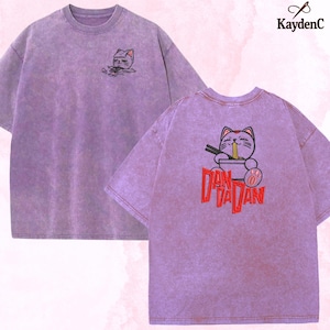 Könnte beinhalten: Ein lila T-Shirt mit einem verwaschenen Effekt. Auf der Vorderseite befindet sich eine kleine Katzen-Grafik, während die Rückseite ein größeres Katzendesign mit Nudeln und dem roten Text "Dan Dan" zeigt. Das KaydenC-Logo ist sichtbar.