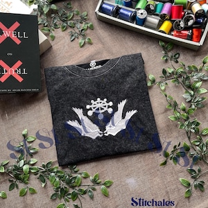 以下が含まれることがあります： チャコールグレーのアシッドウォッシュ加工Tシャツ。中央の車輪のシンボルと、その両側に配置された2枚の翼が白の刺繍で施されています。クルーネックのTシャツが、裁縫用の糸や本と共に茶色の布の上に置かれています。