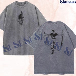 Peut inclure: Un t-shirt gris anthracite délavé minéral avec un graphisme au trait noir représentant un personnage avec une épée sur la poitrine et un graphisme plus grand du même personnage tenant deux épées au dos, avec le texte Stitchales dans le coin.