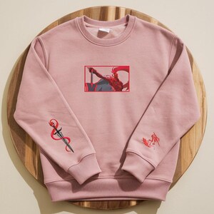 Puede incluir: Sudadera rosa empolvado con bordados. La parte delantera presenta una ilustraci&oacute;n enmarcada de una figura con una espada. La manga izquierda tiene un dise&ntilde;o de espada y la manga derecha un peque&ntilde;o dise&ntilde;o de figura. La sudadera es de material suave.