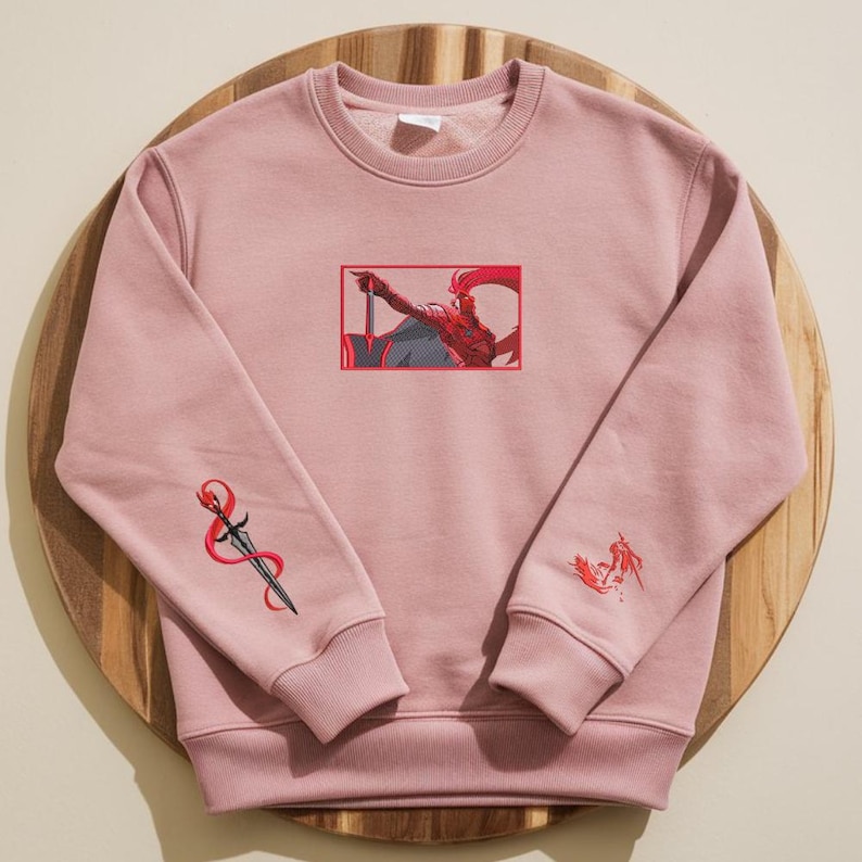 Puede incluir: Sudadera rosa empolvado con bordados. La parte delantera presenta una ilustraci&oacute;n enmarcada de una figura con una espada. La manga izquierda tiene un dise&ntilde;o de espada y la manga derecha un peque&ntilde;o dise&ntilde;o de figura. La sudadera es de material suave.