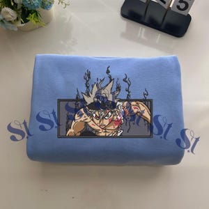 May include: A light blue sweatshirt featuring a rectangular embroidered graphic of an anime character with spiky hair, a black headband, and red eyes, surrounded by dark energy wisps. The character is shown from the chest up in a dynamic pose.
