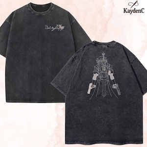 Peut inclure: T-shirts gris foncé avec un aspect vintage délavé. L'un des t-shirts présente le texte "Devil May Cry" avec un graphisme sur le devant. L'autre affiche une illustration détaillée d'un personnage tenant deux armes à feu au dos. La marque "KaydenC" est visible.