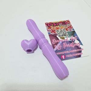 Puede incluir: Una herramienta impresa en 3D de color lila claro con un extremo en forma de corazón y un mango largo y texturizado. La herramienta está junto a una tarjeta de visita con el texto "Diamond Art 3D Printing".