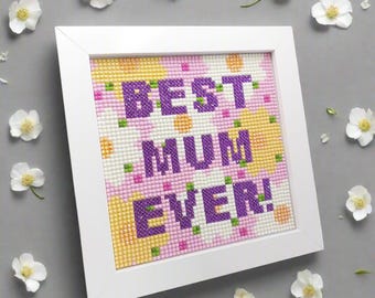 May include: A framed cross-stitch artwork with the text "BEST MUM EVER!" in purple. The background features a floral pattern in yellow, pink, and white. White flowers are scattered around the frame on a gray surface.