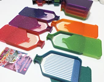 May include: A collection of colorful, plastic diamond painting trays in various colors, including red, purple, green, and blue. Each tray has a lid and a sloped interior for organizing small craft items. A business card with a QR code is also visible.