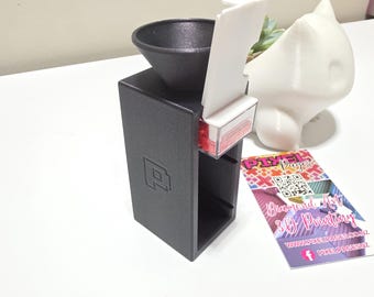 May include: A black 3D-printed dispenser with a black funnel, white lever, and clear container holding red objects. A white figurine and a business card with the text "Pixel Pages" are also visible.