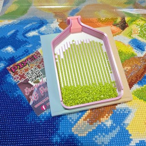May include: A pink diamond painting tray filled with light green diamond-shaped beads. The tray sits on a light blue and beige surface, with a partially visible diamond painting in the background. A business card with the text "Pixel Pages" is also visible.