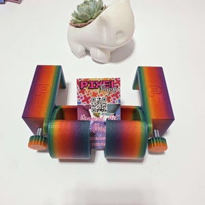 May include: A multi-coloured 3D-printed phone stand with adjustable knobs. A small white hippo-shaped planter holds a succulent. A business card with the text "Pixel Pages" is in the stand.