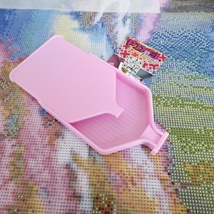 May include: A light pink diamond painting tray with a lid. The tray has a rectangular shape with a spout for easy pouring. A small card with the text "Pixel Pages" is next to the tray.