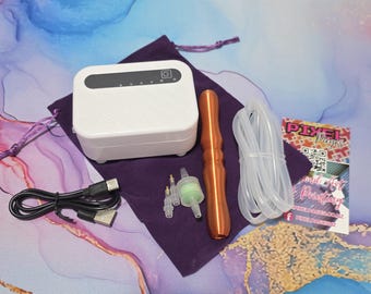 May include: A white rectangular device with a power button and indicator lights, a copper-colored wand, clear tubing, and a USB cable. Accessories include small nozzles and a green filter. A purple drawstring bag and a business card are also present.