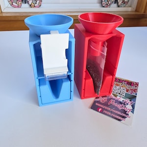 Puede incluir: Dos dispensadores impresos en 3D, uno azul y otro rojo, con tapas en forma de embudo y cuerpos rectangulares. El dispensador azul tiene un componente blanco. El dispensador rojo contiene una bolsa transparente llena de cuentas oscuras.