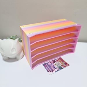 Puede incluir: Un organizador rosa y arcoíris de seis niveles. La parte superior del organizador tiene un degradado de amarillo, naranja y rosa. Una figura decorativa blanca de un personaje de dibujos animados y una tarjeta de visita están junto al organizador.
