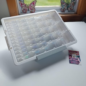 Puede incluir: Un contenedor de almacenamiento de plástico transparente con múltiples compartimentos pequeños, ideal para organizar objetos pequeños. El contenedor tiene un marco blanco y un asa gris. Una tarjeta de visita está a la derecha. Dos decoraciones de ventana de mariposa están en el fondo.