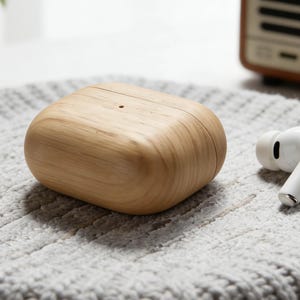 May include: A wooden effect case for wireless earbuds rests on a grey knitted surface. Two white wireless earbuds are next to the case. The case has a smooth, rounded shape and a natural wood grain appearance. The earbuds have a sleek, modern design.