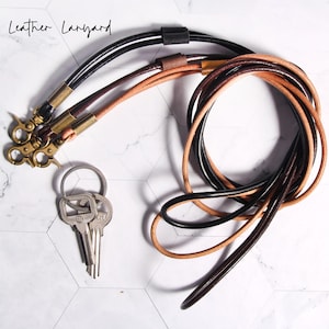 May include: Leather lanyards in black, brown, and tan leather with brass-coloured clasps. A set of keys is attached to one of the lanyards. The lanyards are arranged on a white and grey hexagonal tile surface.