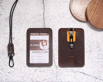 May include: Two dark brown leather ID card holders with a black lanyard. One holder displays a press pass with the name Amanda Clark, while the other features a black metal clip attached to a yellow leather accent on the back of the card case.
