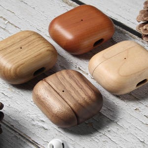 Op de afbeelding: Vier houten AirPod-hoesjes in verschillende bruin- en beigetinten. De vierkante hoesjes hebben afgeronde randen en een klein gaatje aan de zijkant. De houtnerf is zichtbaar op elk hoesje, geplaatst op een wit houten oppervlak. Elk hoesje is ongeveer 5 cm groot.