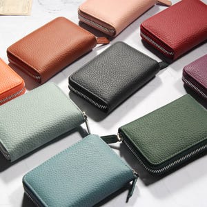 Puede incluir: Una colección de carteras con cremallera de colores, en varios tonos como naranja, marrón, negro, verde y morado. Las carteras tienen una superficie texturizada y cremalleras plateadas. La disposición sugiere una exhibición de accesorios.