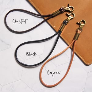 May include: Three leather wrist straps in chestnut, black, and cognac colors, each with a brass-colored clasp. The straps are looped and lie on a white and gray hexagonal tile surface. The word "Chestnut", "Black", and "Cognac" are written on the surface.