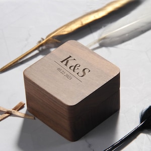May include: A square wooden box with the engraved initials "K&S" and the date "05.22.2023" on the lid. The box is surrounded by decorative feathers and ribbons. The box is made of a light brown wood.