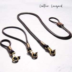 Könnte beinhalten: Vier braune Leder-Lanyards mit goldfarbenen Verschlüssen und Schlaufen. Die Lanyards sind unterschiedlich lang, das längste ist etwa 76 cm lang. Der Text "Leather Lanyard" steht in Kursivschrift oben.