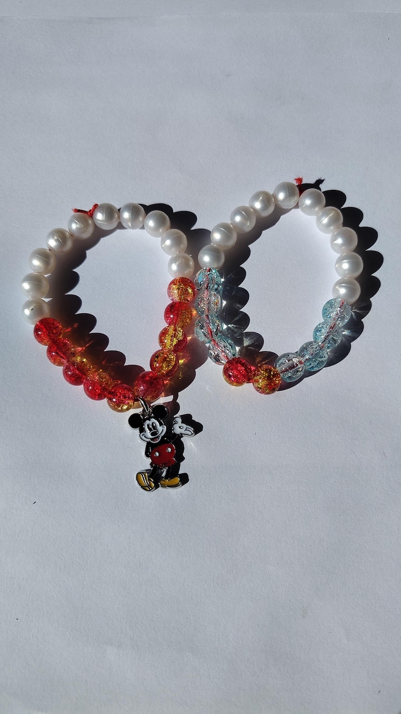 K&ouml;nnte beinhalten: Zwei Armb&auml;nder mit Perlen. Jedes Armband hat wei&szlig;e Perlen und farbige Perlen in Rot, Orange und Blau. Ein Armband hat einen Mickey Mouse-Anh&auml;nger. Die Armb&auml;nder liegen auf einer wei&szlig;en Oberfl&auml;che.