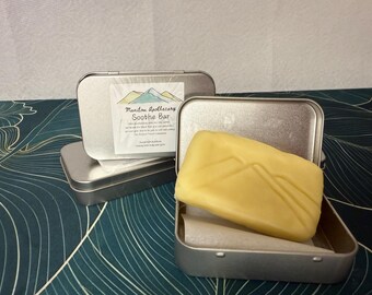 May include: A light yellow Soothe Bar in an open silver tin, with a second tin and a closed tin in the background. The bar has a mountain design. The label reads "Manitou Apothecary Soothe Bar". The background is a teal and white leaf pattern.